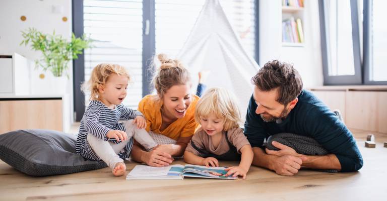 Jong gezin met twee kleine kinderen binnenshuis in de slaapkamer die een boek lezen.